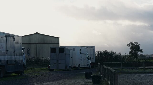 A line of white horse trailers and transport trucks parked near a large metal barn. Professional equestrian transport equipment in a rural countryside setting under a cloudy sky.