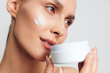 Closeup of young woman applying moisturizing cream on cheek while holding white container isolated on light background with soft natural light and healthy skin care concept.