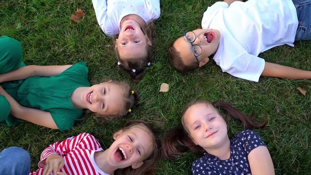 Top view of happy little children lie in circle on grass in summer park look at camera and laugh. Girls and boys play outdoors. Kids leisure