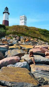 View of woman crocheting on rocky shore with coastal lighthouse. Long shot of a woman sitting on grey and brown riprap rocks crocheting near a historic white and red lighthouse.