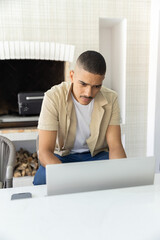 Naklejka premium Man in his 30s sitting at home using laptop, smartphone left, wearing beige shirt, fireplace behind