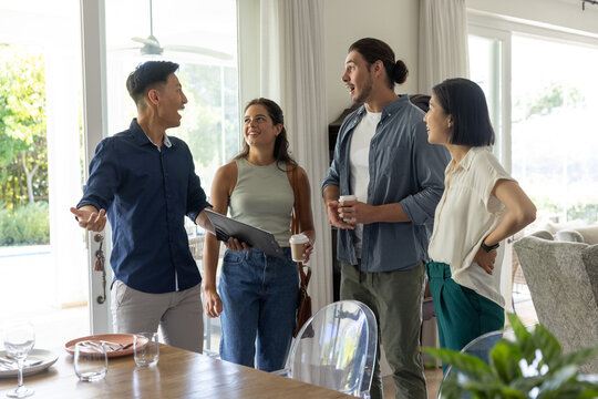 Diverse friends standing and chatting around dining table at home, holding tablet and coffee cups