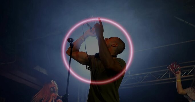 Singing lead vocalist in dark green tee lit by pink-halo on stage, holding mic, phones, tambourine