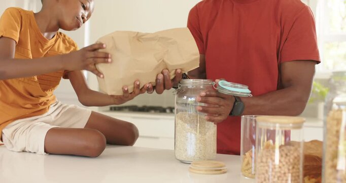 African American father and child tipping brown bag, steadying blue-lidded jar to store oats