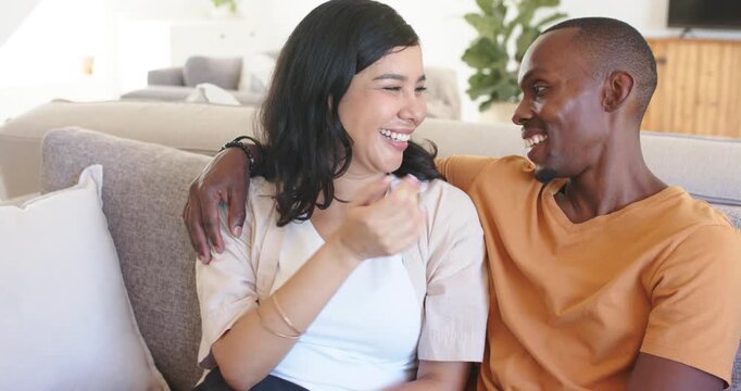 Couple sitting on gray sofa in bright living room, man leaning, draping arm, woman laughing, posing