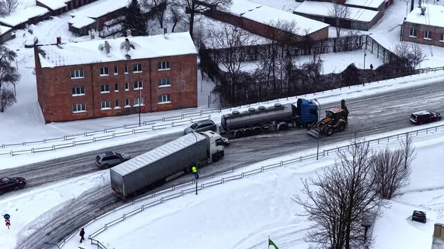 Aerial winter scene of a jackknifed semi trailer blocking an icy city road as cars pass and a recovery vehicle assists after a traffic accident in snowy conditions.