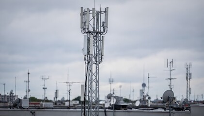 Medium shot of a rooftop antenna farm with a tall broadcast antenna in sharp focus against a blurred background of other antennas under a cloudy sky.
