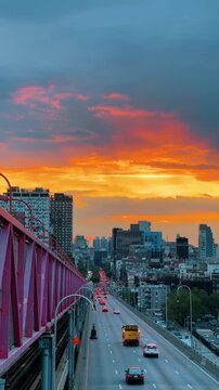 Sunset over urban highway with heavy traffic and bridge. Vibrant orange sunset over city road with cars and pink metal bridge railing in the foreground.
