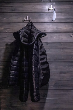 Black shiny jacket hanging alone on clothes hanger in dark wooden hallway interior. Minimalist coat rack scene with glossy outerwear in moody home entrance.