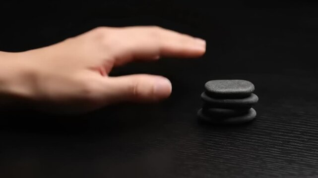 Hand stacking smooth dark stones on a black surface for balance and meditation.