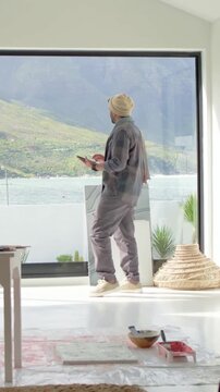 Vertical video: Comparing tablet and sea view, Indian artist in beanie planning canvases at studio