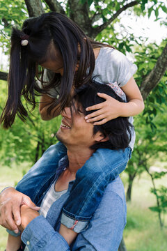 A father carries his daughter on his shoulders as she playfully covers his eyes with her hands. They are outdoors surrounded by trees and greenery.