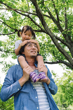 A father carries his young daughter on his shoulders as they enjoy a sunny day outdoors amidst lush green trees.
