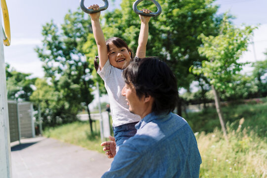 A father watches his daughter swing on monkey bars at a park. The girl smiles joyfully as she hangs from the rings, enjoying outdoor playtime with her dad.