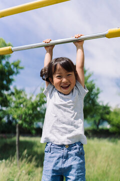 A joyful young child hangs from a monkey bar, smiling broadly outdoors on a sunny day.