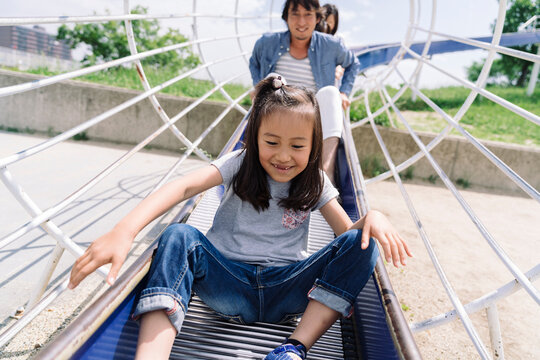 A young girl slides down a playground structure with her parents behind her, enjoying a fun day outdoors.