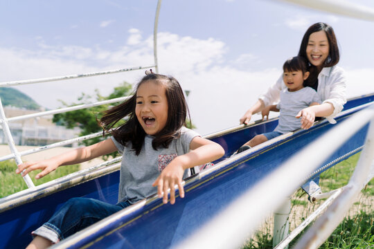 A mother and her two children enjoy a sunny day sliding down a blue slide together, sharing laughter and fun.