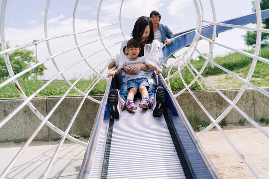 A family enjoys a sunny day at the park, with parents and children sliding down a playground slide together.