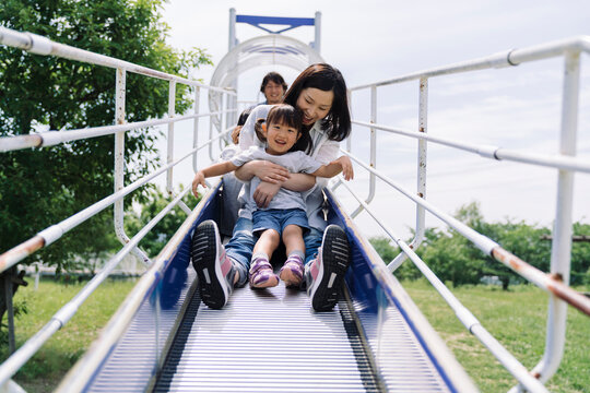 A family enjoys a fun day at the park, with a mother and her children sliding down a playground structure together.