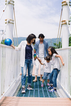 A family walks together on a suspension bridge, holding hands and enjoying a day outdoors. The parents guide their two young daughters across the walkway.