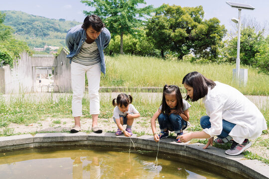 A family enjoys a sunny day outdoors, with parents and two young children gathered around a water feature, observing and interacting with the water.