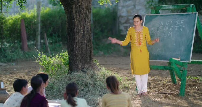 Indian Young adult gen z female tutor standing teach English to villager girl boy outdoor rural area. Happy child group sitting on mat look board learn do study new day time outside ground place