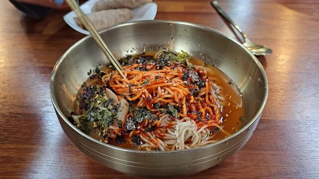Chopsticks stir spicy red gochujang buckwheat noodles topped with dried seaweed and sesame seeds in traditional Korean steel bowl served with bossam wrap
