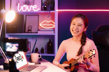 Fototapeta premium Host channel in musician of young beautiful Asian playing ukulele along singing, broadcasting on social media channel by phone at neon colorful light modern studio recording by camera. Stratagem..