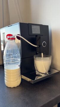 Automatic coffee machine frothing milk into a glass. Close-up of hot milk foam flowing from the dispenser nozzle, using a milk tube connected to a bottle. Modern home barista equipment.