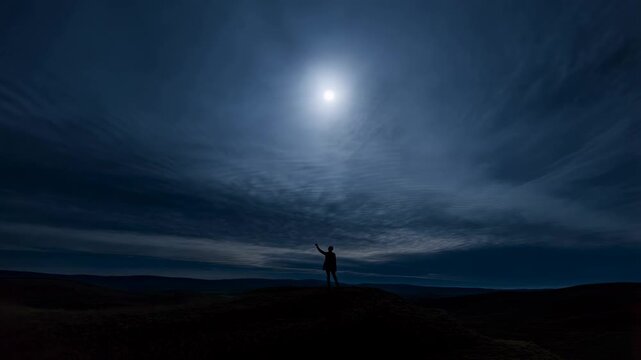 Slow pan of lone person raising arm on hill under moonlit cloudy sky in remote rolling landscape at night