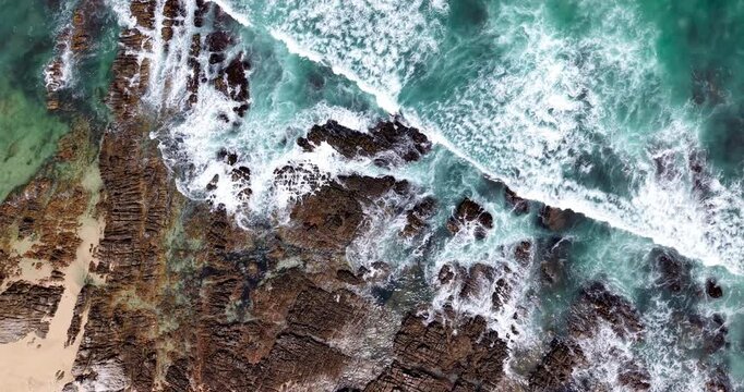 Drone top-down aerial view of turquoise ocean waves breaking over rugged rock formations along a sandy shoreline. White sea foam washes across the reef and rocky coastline, revealing natural patterns 