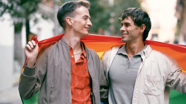 Happy gay couple lovingly looking at each other while wrapped in a rainbow pride flag. Authentic portrait of two men celebrating their love and identity