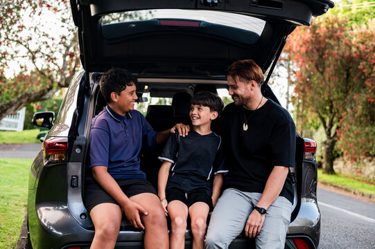 Father sitting beside two boys making fun in an open boot of parked car
