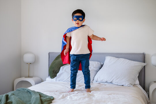 Young Asian boy wearing a mask and cape jumps joyfully on a bed in his room