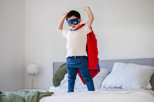 Asian boy in a superhero costume stands on a bed with arms raised mid air
