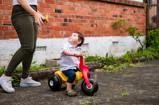 Mum standing beside toddler son riding his tricycle in the backyard
