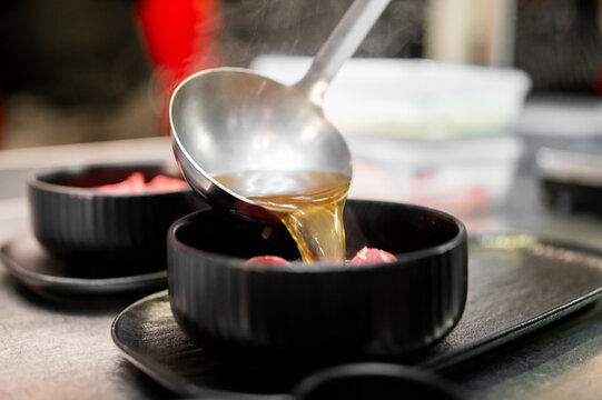 A chef's ladle pours steaming hot, golden broth into black ceramic bowls filled with thinly sliced raw meat, captured in a professional kitchen setting with a shallow depth of field
