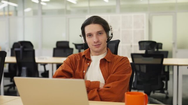 Young male operator wearing headset while working in office. Sitting at desk and consulting customer through laptop. Portrait with smile in call center