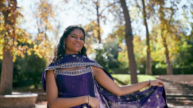 Beautiful young indian woman with a bindi smiling and dancing gracefully in a park. She wears a traditional purple sari during a celebration