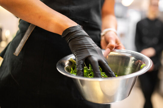 A chef wearing black nitrile gloves and a black apron mixes a fresh green salad in a stainless steel bowl in a professional kitchen