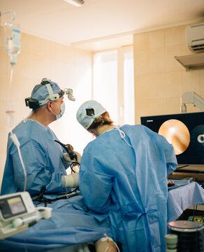 Medical team consisting of two surgeons operating on a patient using specialized endoscopic equipment and monitoring internal views on a screen in a hospital environment