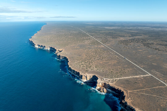 Aerial view of dramatic limestone cliffs dropping to deep blue ocean beside a straight outback road
