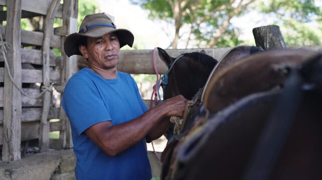 Latin man working in a rustic wooden stable. Old cowboy wearing a hat preparing a beautiful dark brown horse for a ride on a ranch