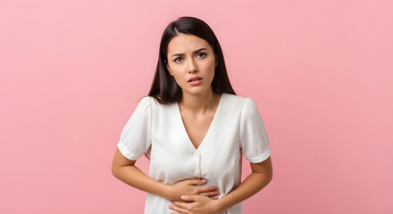 Fototapeta premium Woman with dark hair wearing white shirt looks concerned, holding her stomach in discomfort or pain on pink background