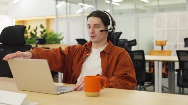 Call center operator taking off headset after long work. Sitting at desk with orange cup with coffee. Closing his laptop after work. Getting relief and taking the breathe