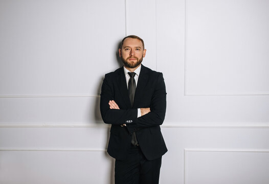 Photograph of a stylish, mature businessman in a black suit and tie standing against a white wall in his office. Business concept.