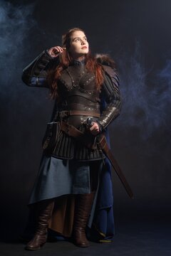 A warrior woman stands confidently amidst swirling smoke, clad in detailed armor and a flowing blue cloak.