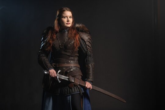 A fierce warrior woman stands poised with a sword against a dark backdrop, clad in armor and ready for battle.