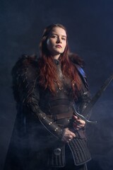 Obraz premium A fierce warrior woman with fiery hair and a sword stands ready against a smoky, dark background.