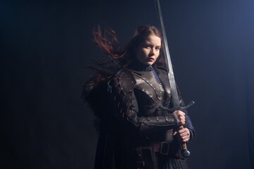 A fierce warrior woman stands poised with a sword, clad in dark armor against a dramatic backdrop. © Fotokvadrat
